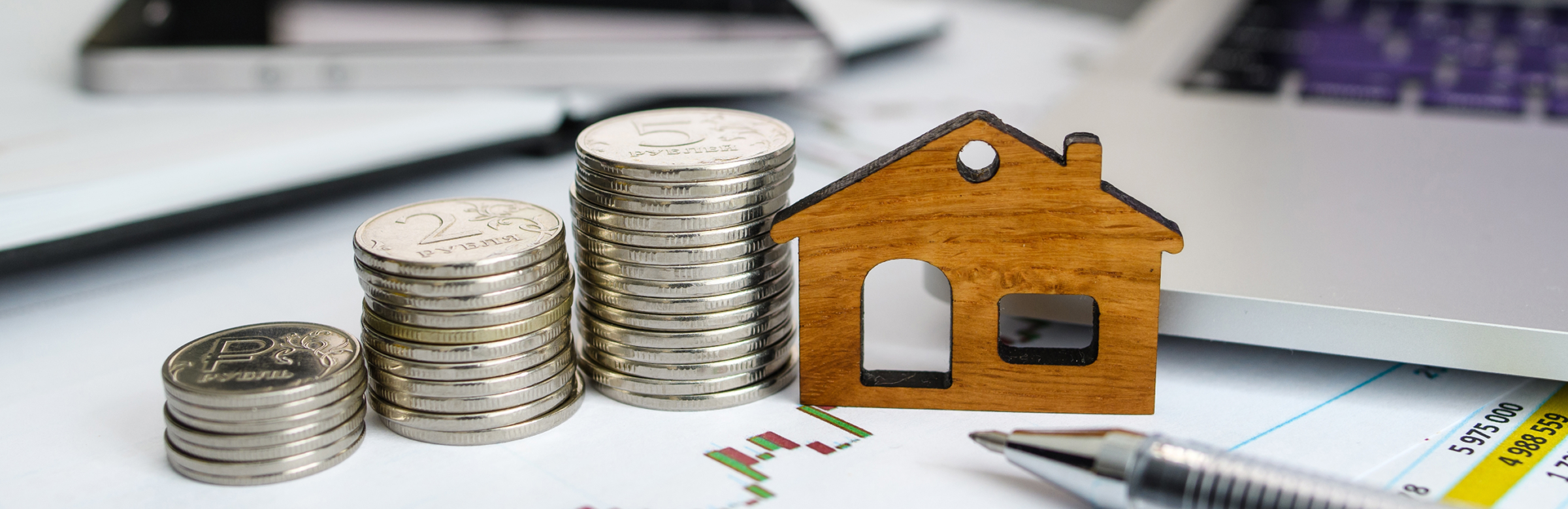 Piles de pièces d'argent à côté d'une maquette de maison en bois sur un bureau avec des documents financiers et un ordinateur portable, représentant le financement immobilier et la planification hypothécaire.
