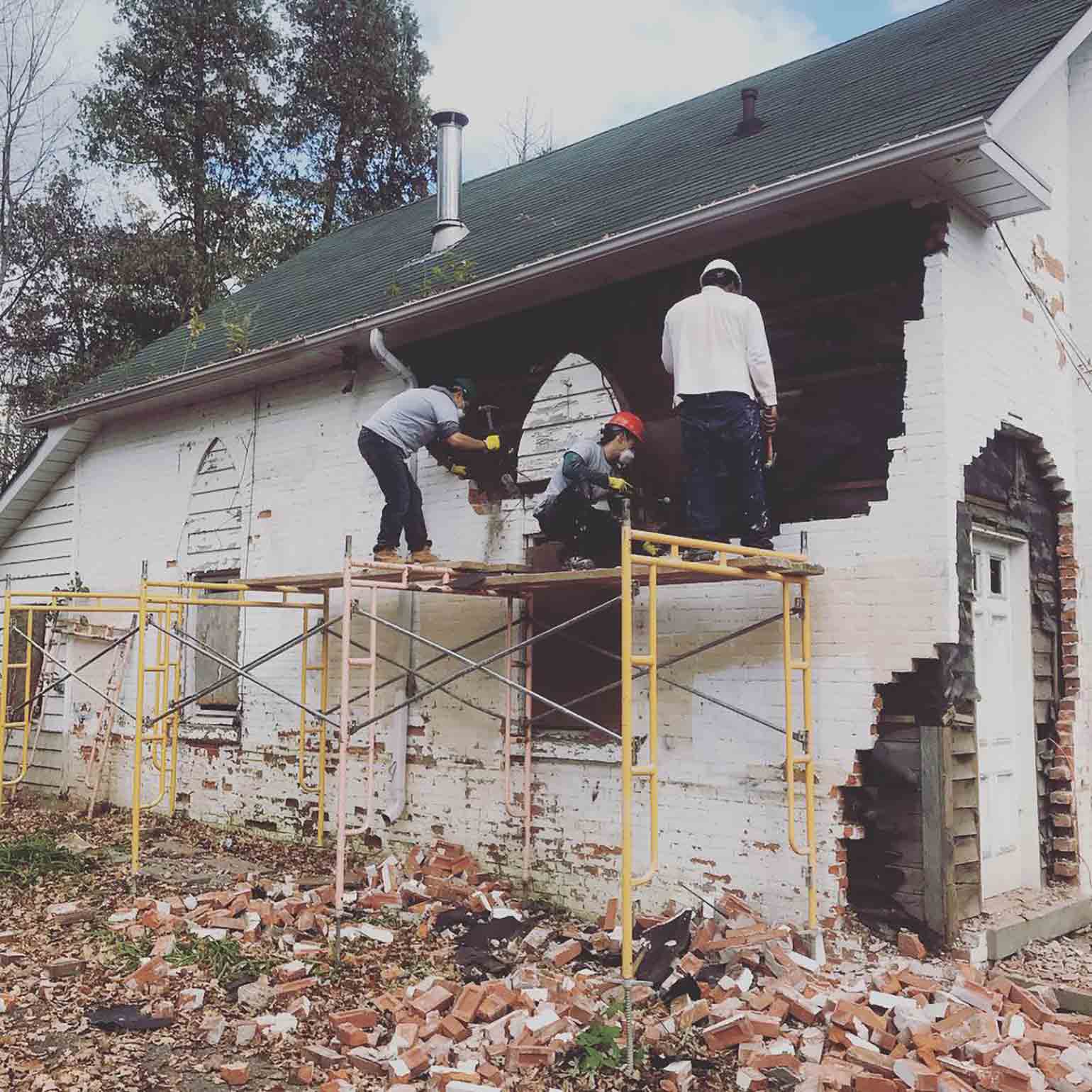 Un constructeur et un apprenti transportent du bois sur un chantier de rénovation. Trois travailleurs aident à démolir un mur de briques blanches pour rénover une maison destinée à loger des sans-abri.