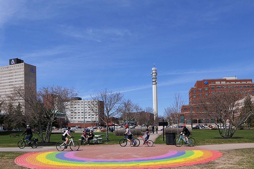  Une place de parc par une journée ensoleillée au centre-ville de Moncton avec des jeunes à vélo et un arc-en-ciel peint sur le trottoir.