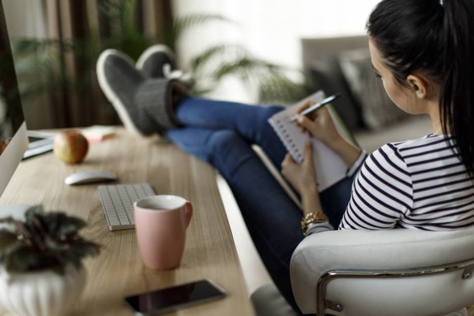 Une femme vue de dos qui écrit dans un carnet, les pieds sur un bureau.