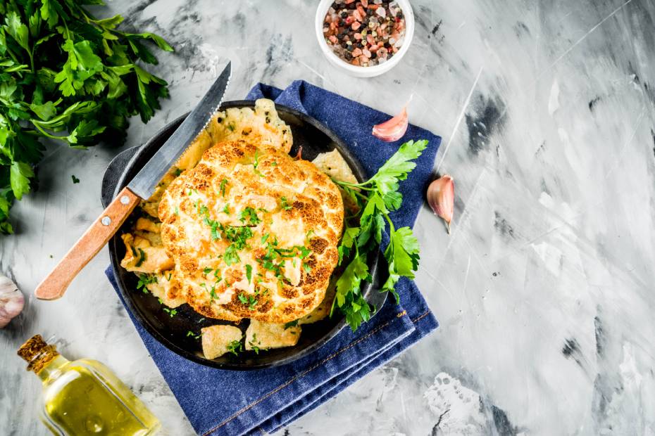 Chou-fleur entier rôti dans une cocotte sur un comptoir en marbre gris et blanc avec des herbes et une bouteille d'huile d'olive.