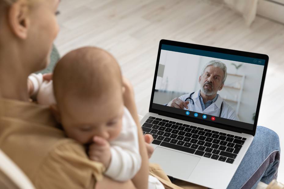 L'image montre une mère tenant son bébé et utilisant un ordinateur portable pour faire un appel vidéo avec un pédiatre.