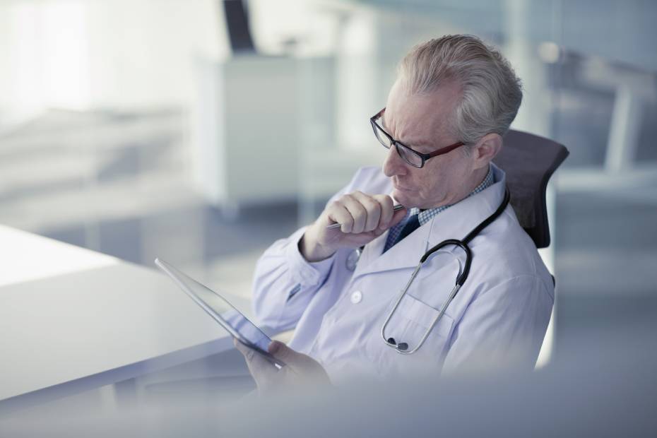 Un médecin d'un certain âge en pleine réflexion regarde sa tablette au bureau.