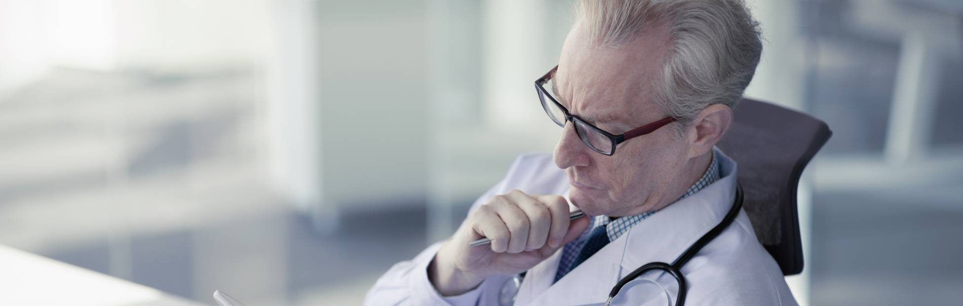 Un médecin d'un certain âge en pleine réflexion regarde sa tablette au bureau.