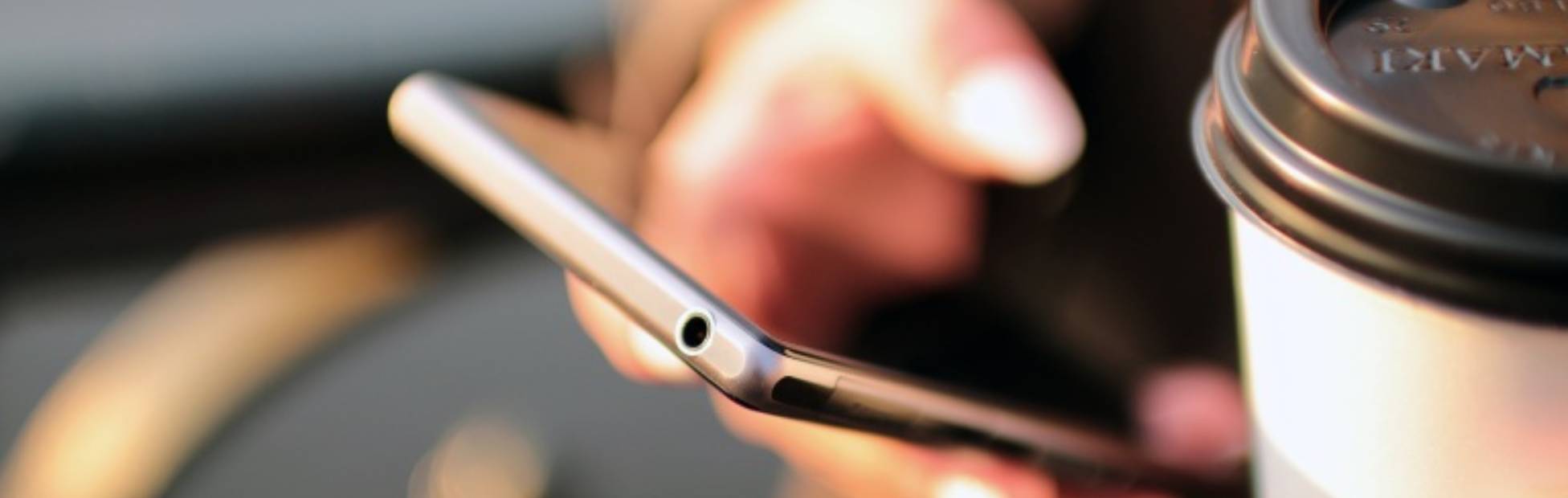 Image d'une femme consultant d'une main son téléphone cellulaire et tenant dans l'autre un café.