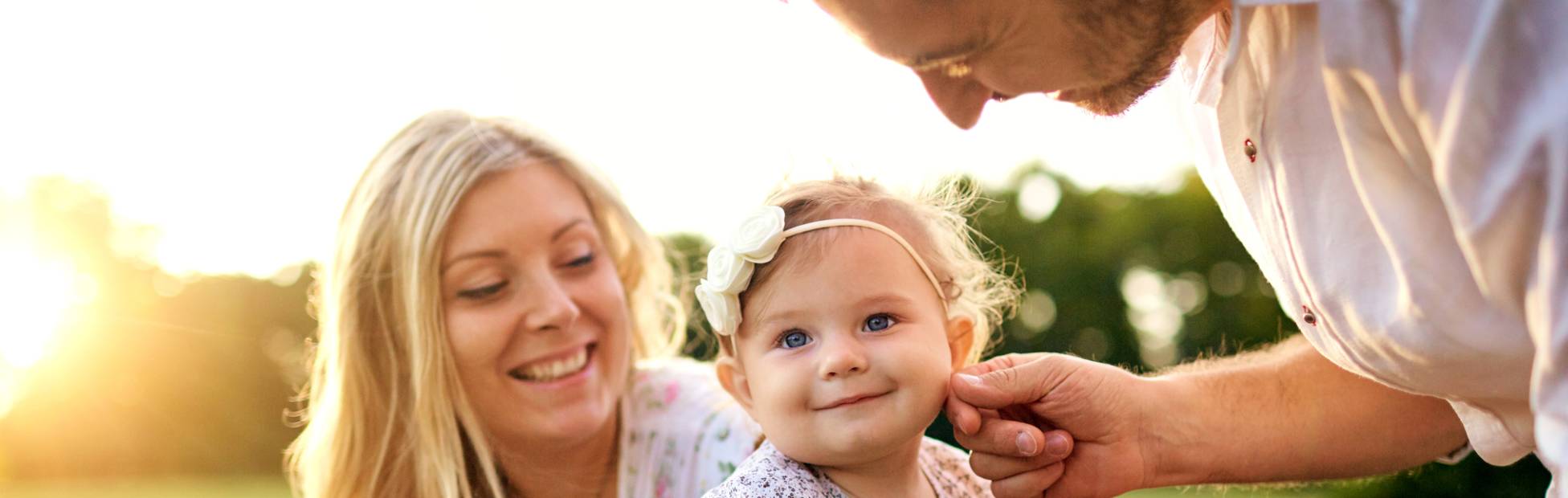 Image d'une jeune famille heureuse installée sur un banc de parc ; le père caresse le visage de la petite fille que tient la mère.