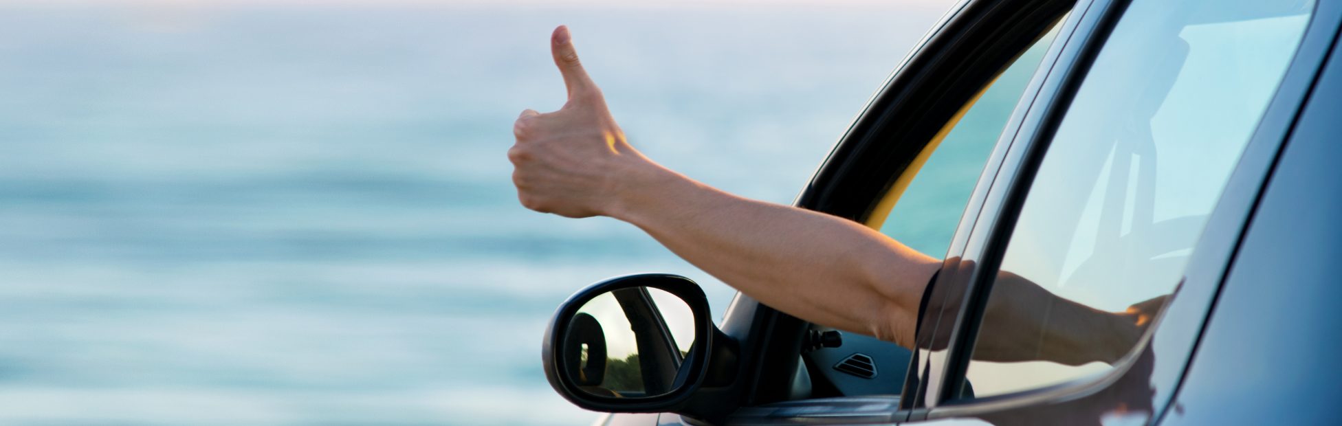 Un conducteur dans une voiture au bord de l'océan sort le bras par sa fenêtre et fait un signe de pouce levé