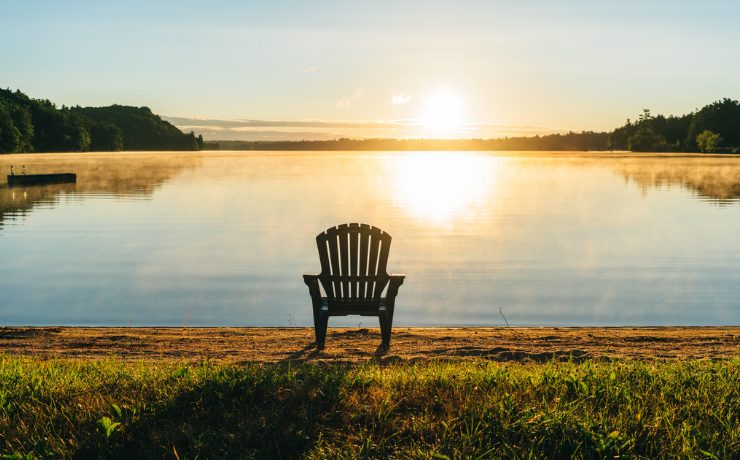 Une chaise Muskoka vide sur une petite plage à côté d'un lac calme au coucher du soleil.