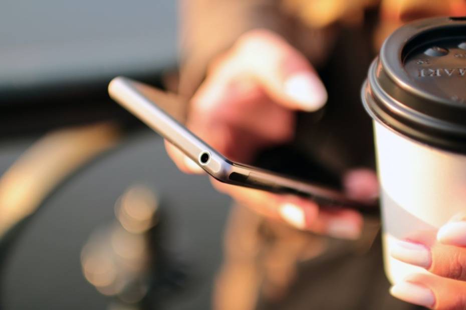 Image d'une femme consultant d'une main son téléphone cellulaire et tenant dans l'autre un café.