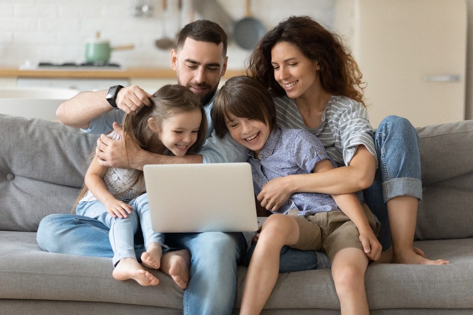 Une jeune famille souriante de quatre personnes assise sur un canapé, regardant un ordinateur portable